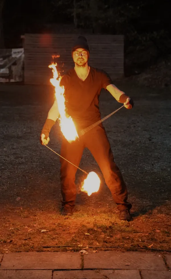 Eine Flammenzunge steht senkrecht vor dem Körper, der zweite Poi kreist tief.
